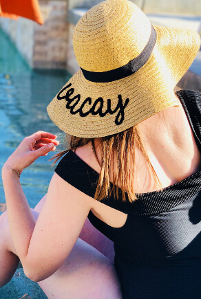 Dáma sedící u bazénu v černých jednodílných plavkách se slaměným kloboukem
